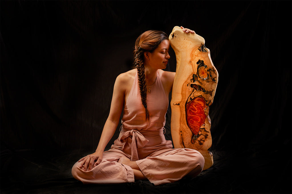 Woman seated on the floor holding a large handcrafted wooden art piece with vibrant red resin inlays against a dark background.
