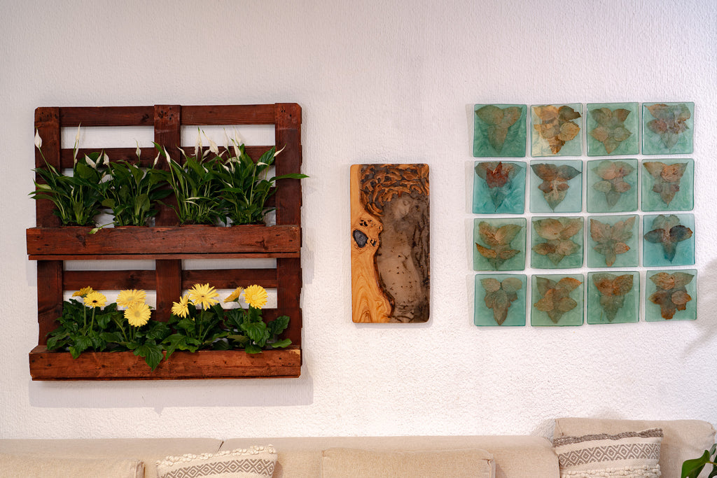 Decorative interior wall featuring a vertical wooden planter with green plants and yellow flowers, a central wood-and-resin art piece, and a grid of sixteen leaf-embedded tiles; beige couch with patterned cushions below.