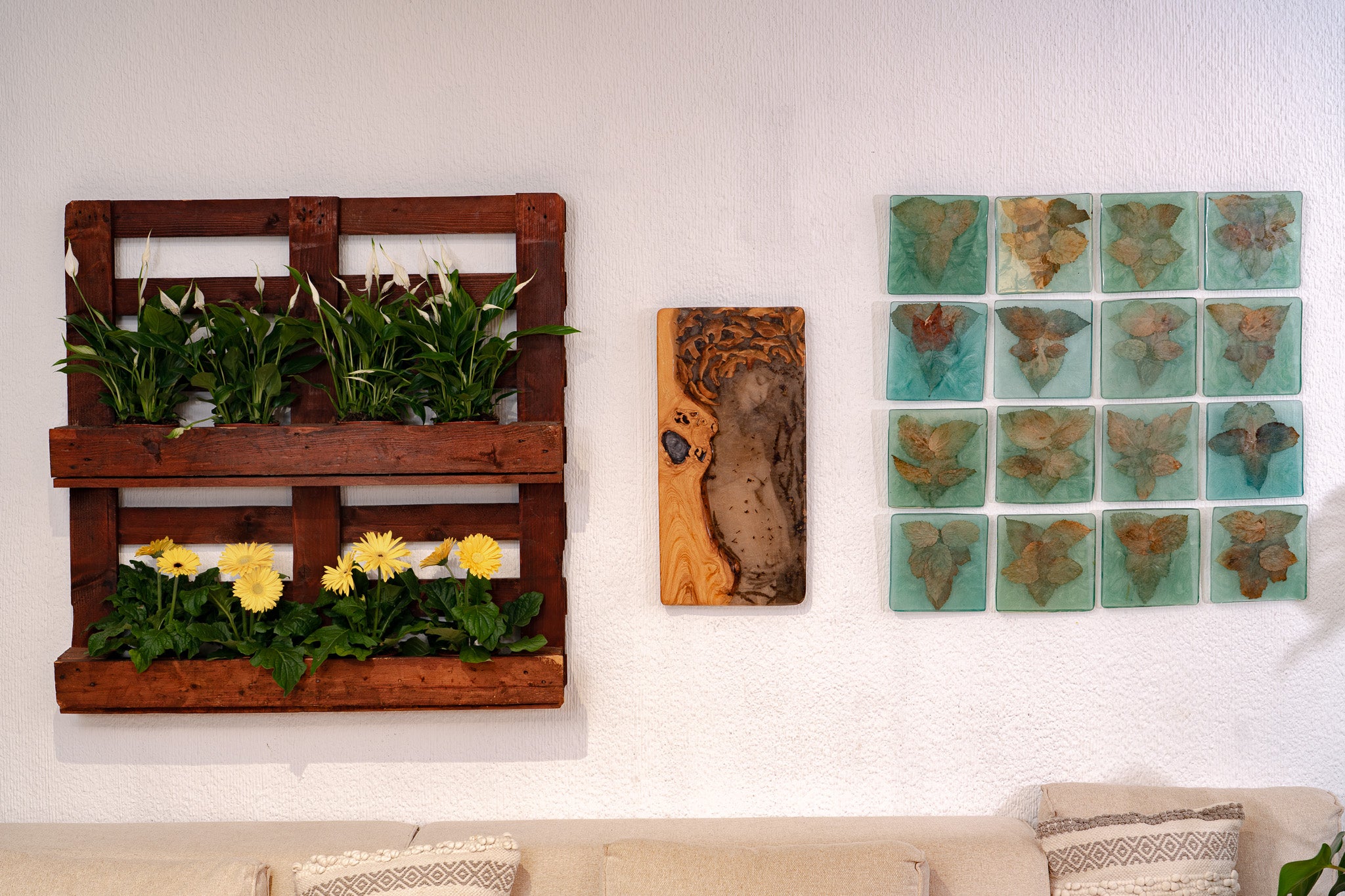 Decorative interior wall featuring a vertical wooden planter with green plants and yellow flowers, a central wood-and-resin art piece, and a grid of sixteen leaf-embedded tiles; beige couch with patterned cushions below.
