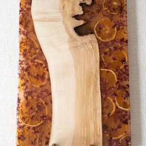 Tabla de servir única con diseño de madera natural y resina translúcida con incrustaciones de rodajas de naranja y pétalos de flores, soportada por asas de latón