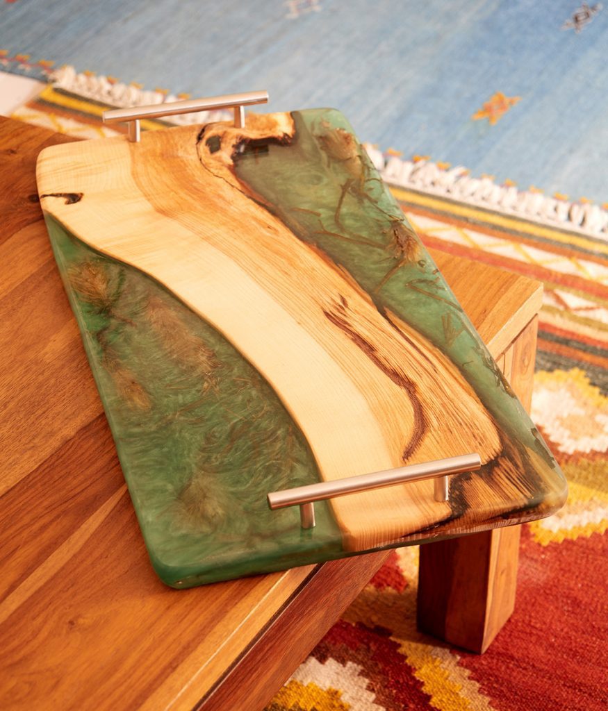 Tabla de servir de madera con resina verde colocada sobre una mesa de madera, con asas de metal plateado.