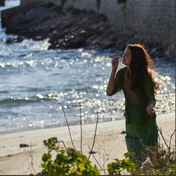 Mujer en vestido verde de pie en la playa mirando al mar, sosteniendo un objeto en su mano, con rocas y olas en el fondo.