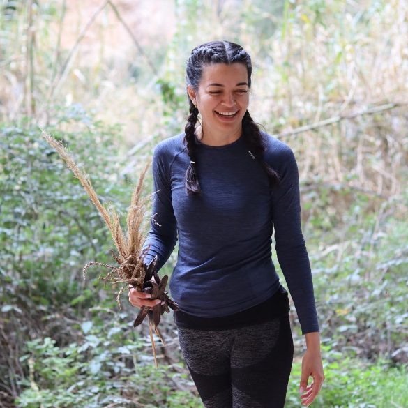 Mujer sonriente en ropa deportiva sosteniendo plantas silvestres en un entorno natural, expresando alegría y conexión con la naturaleza.