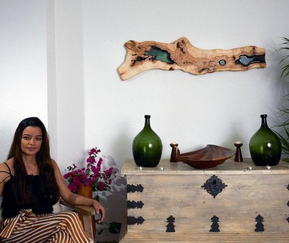 Mujer sentada junto a una cómoda de madera con adornos de botellas verdes, un cuenco decorativo y una pieza de arte de madera en la pared.