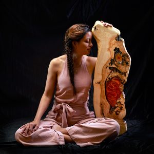La imagen muestra a una mujer en un momento de reflexión junto a una obra de arte en madera y resina, subrayando la belleza y profundidad del arte artesanal